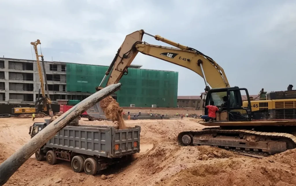 Excavator loading a dump truck beside multi-storey buildings