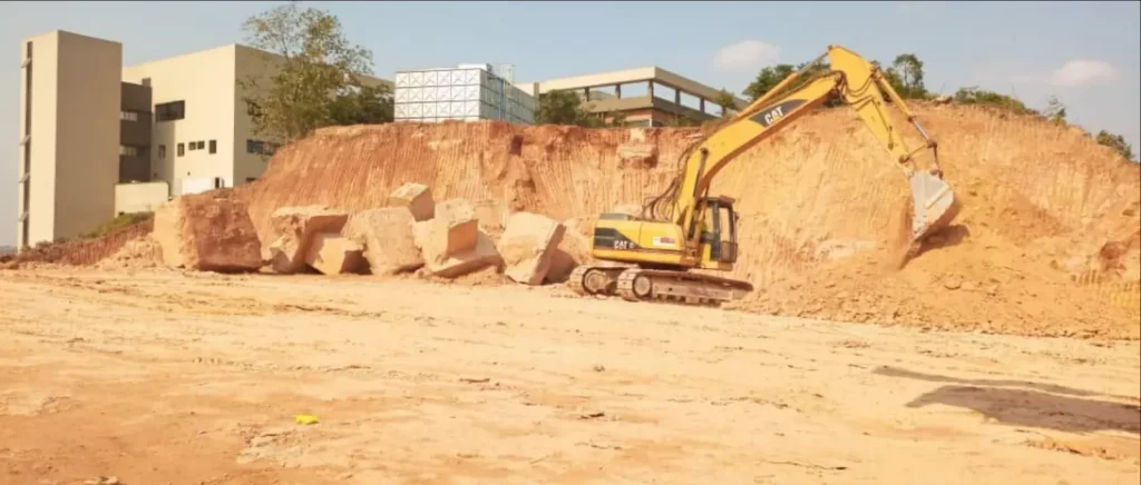Yellow excavator working on a mound of earth and large boulders with buildings behind
