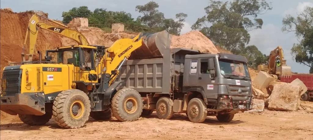 Wheel loader loading a tipper truck beside large boulders and an excavator